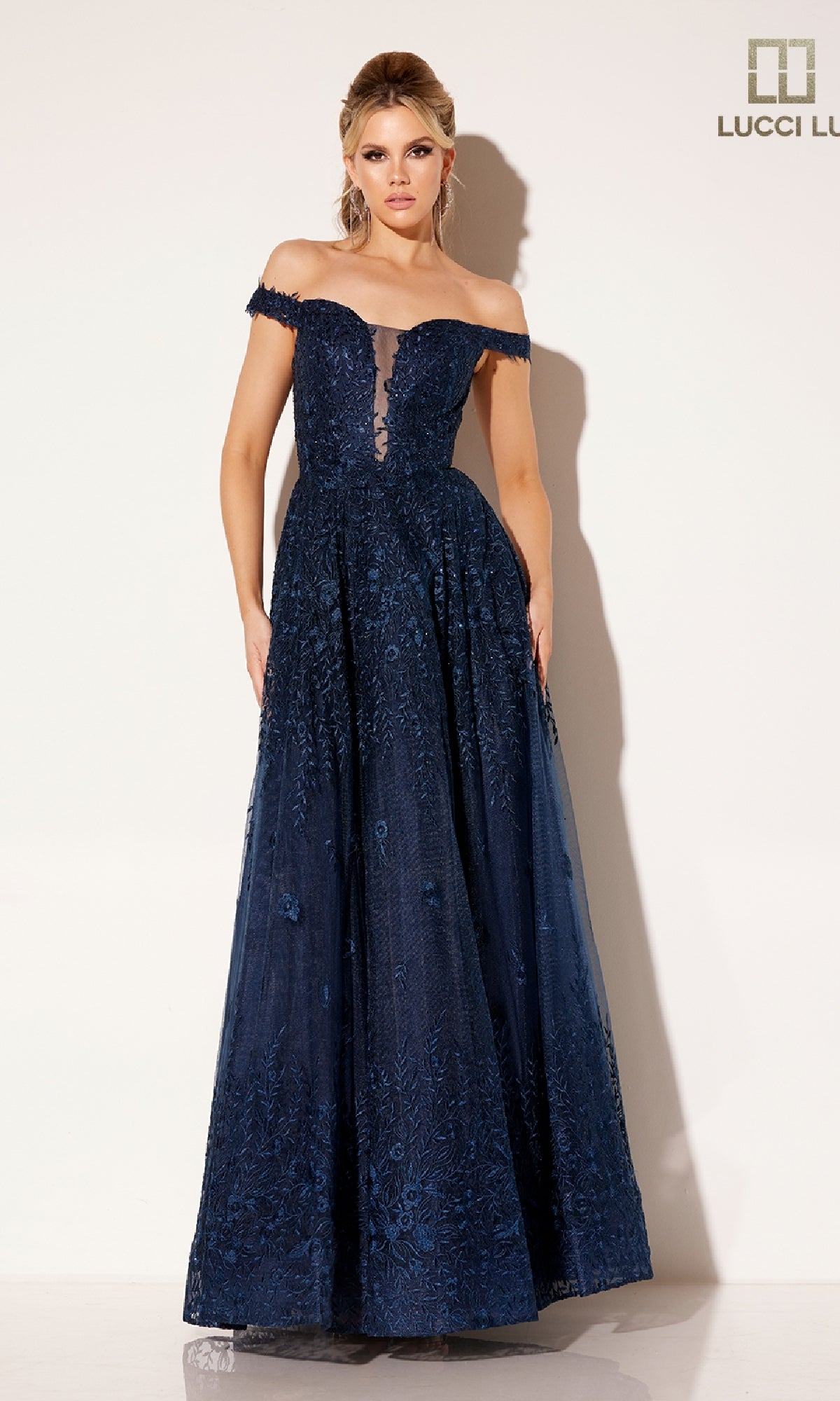 A woman models the Lucci Lu C8090 Long Prom Dress, featuring navy blue lace embroidery, an off-the-shoulder neckline, and a floor-length skirt, set against a plain light background.